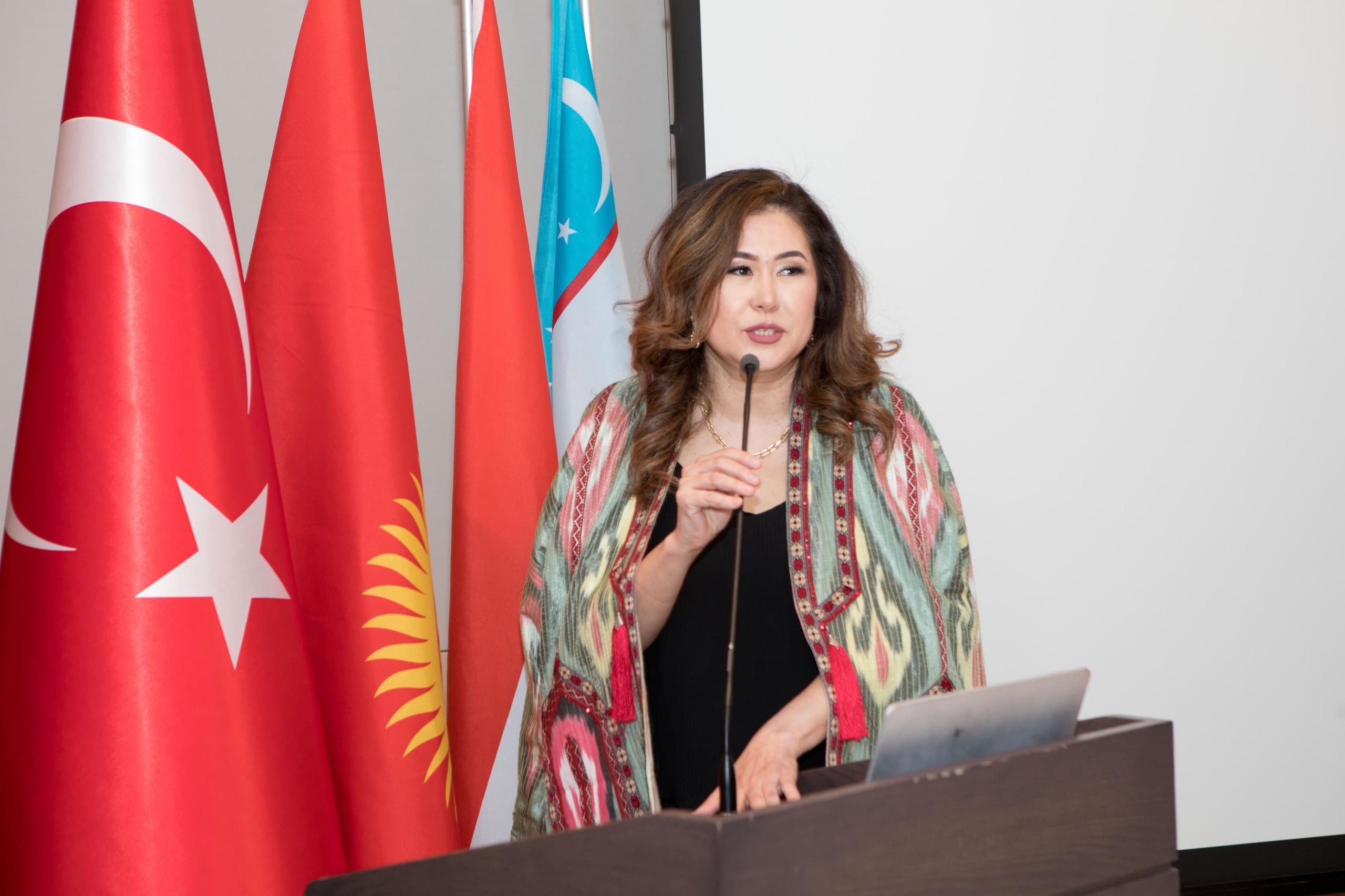 Speaker in an ikat robe addressing guests from a podium beside the flags of Türkiye, Kyrgyzstan and Uzbekistan